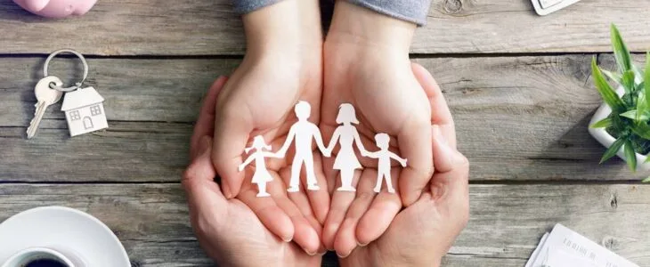 "Four hands cupped around a paper cut-out family, symbolizing family protection, on a wooden desk with a piggy bank and coffee."